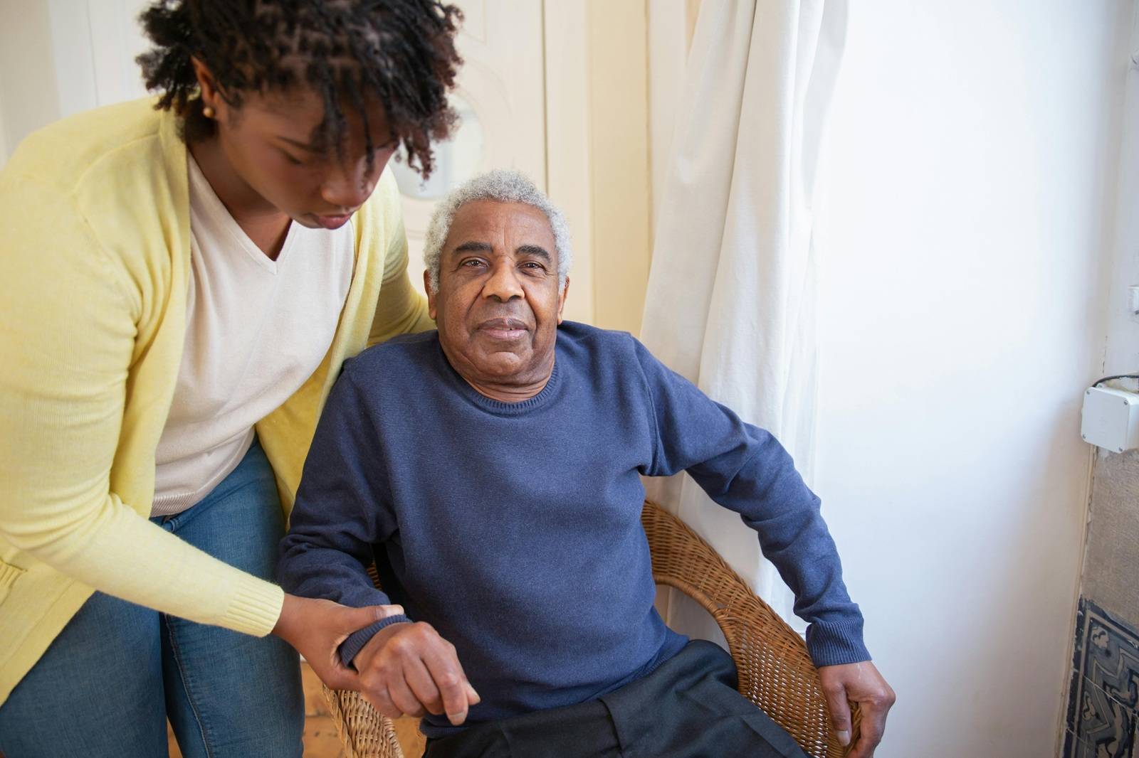 Adult daughter helping her elderly father stand from a chair in a home setting.
